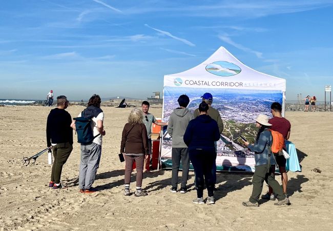 People gathered around a tent labeled Coastal Corridor Alliance on the beach with blue skies above.