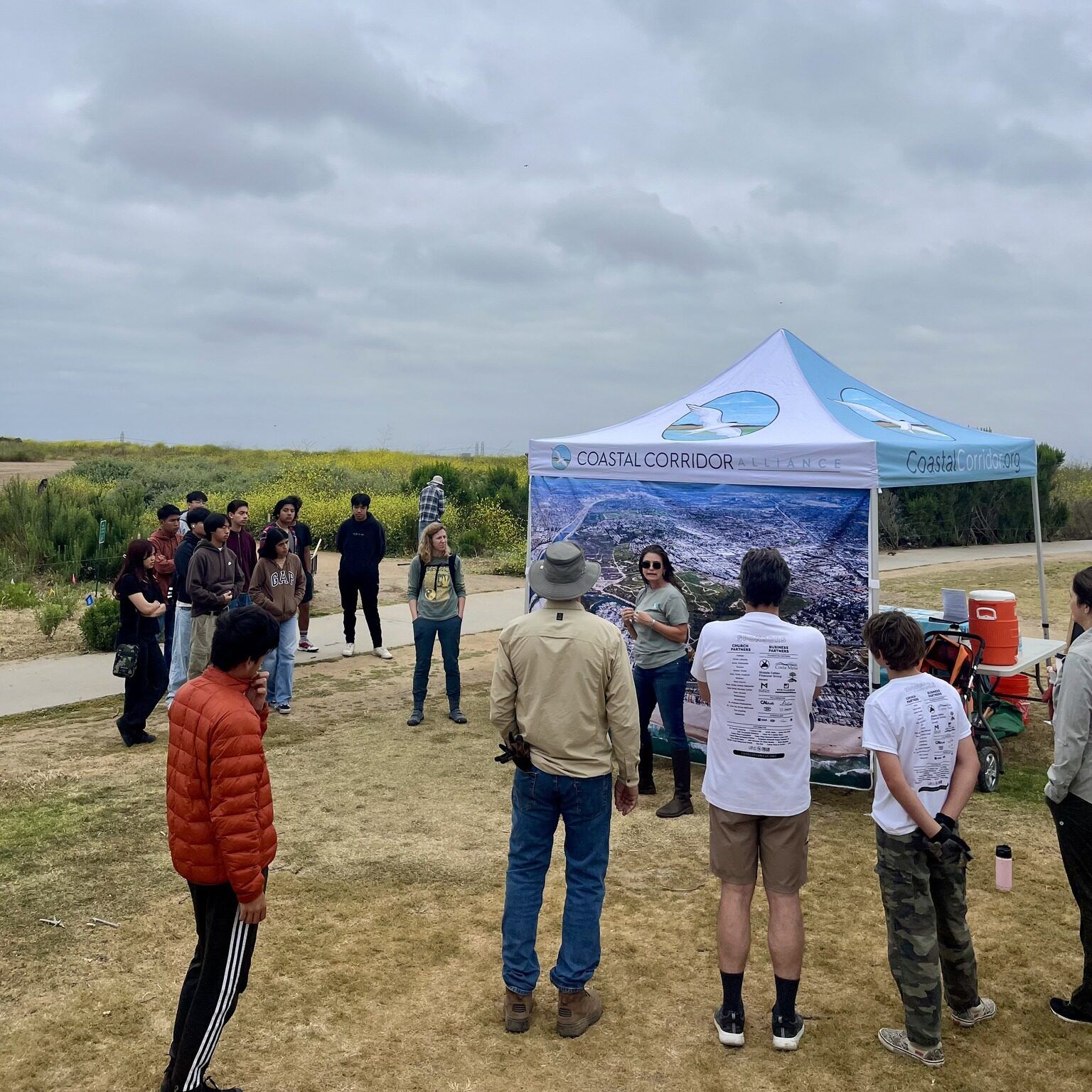 A group of people around a map and canopy listening to a speaker in an outdoor setting with grey clouds above.