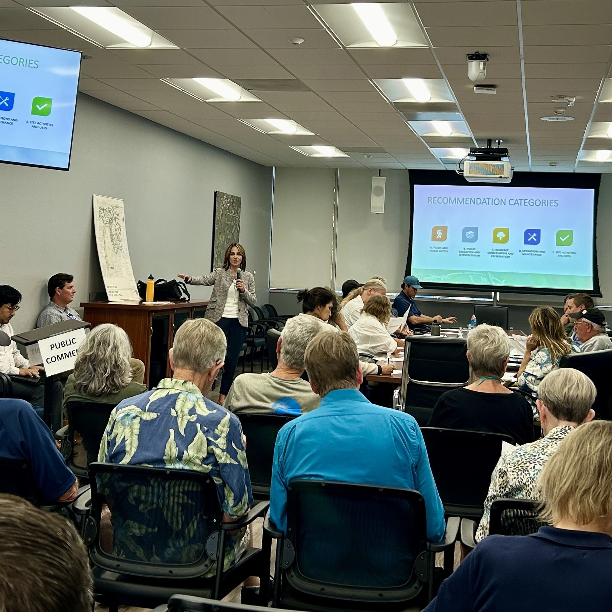 A photograph of people in an audience listening to a presentation with graphic boards and presentation materials around the government room.