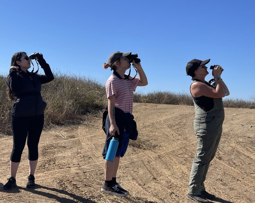 People standing with binoculars on a dirt trail looking at something in the distance.