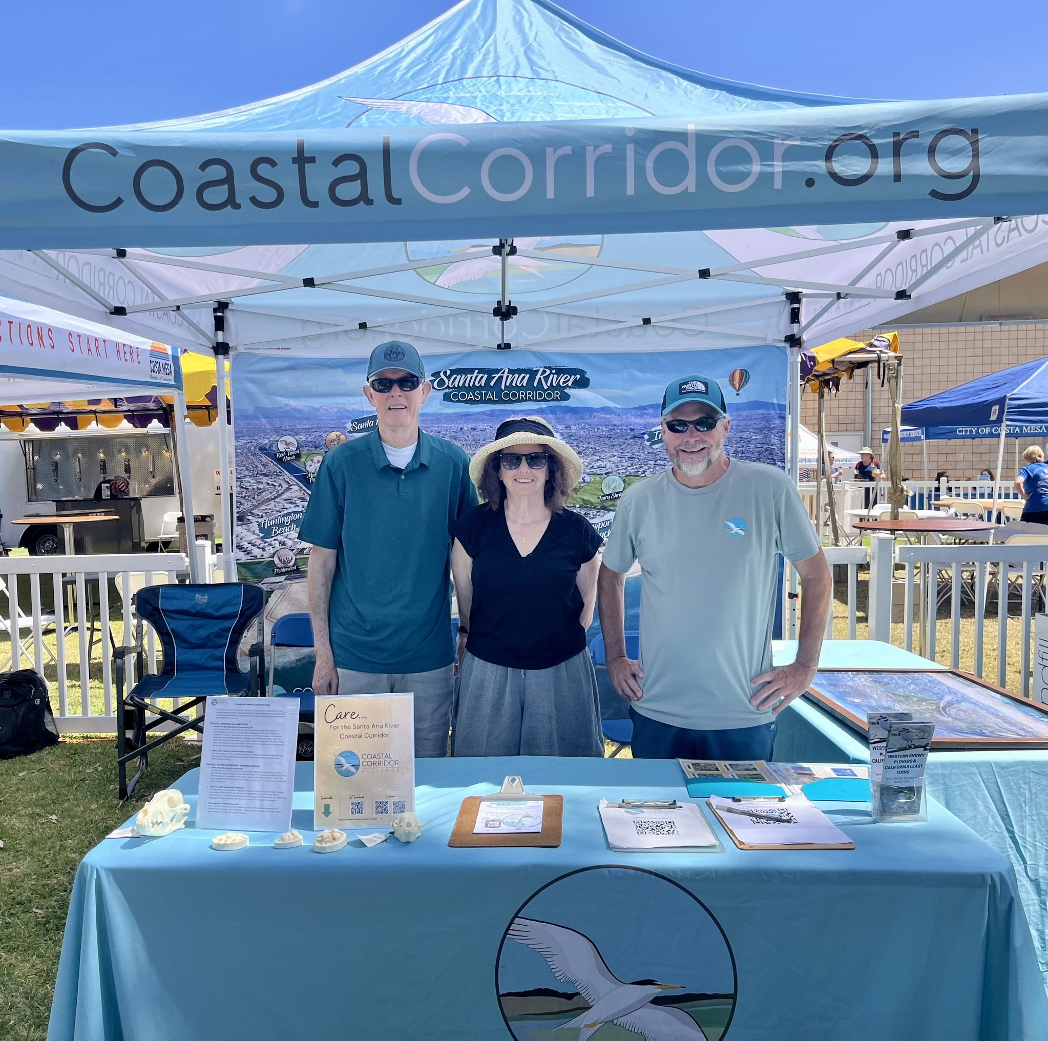 Three people stand at a Coastal Corridor Booth with materials spread over two tables.