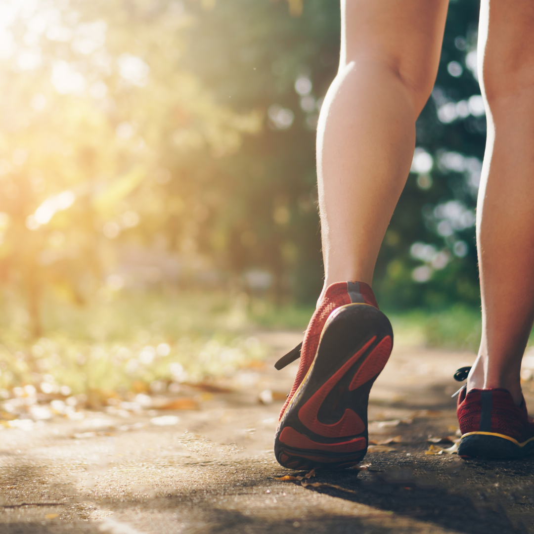 Legs of a woman off on the right with sneakers on. She is standing on a trail outside.