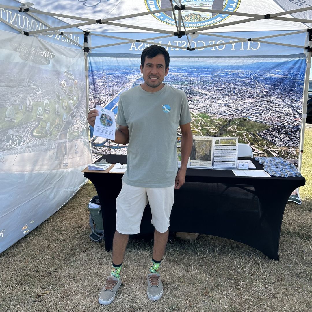 A hispanic man stands in a tent canopy with materials on a table behind him.