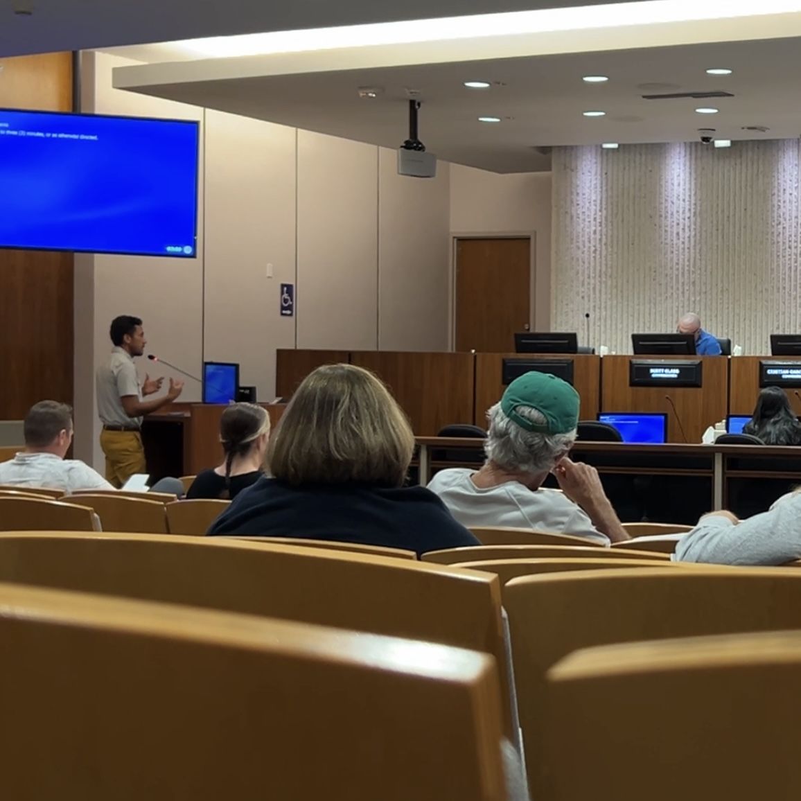 A man stands at a podium speaking to commissioners at the dias with people in the audience.