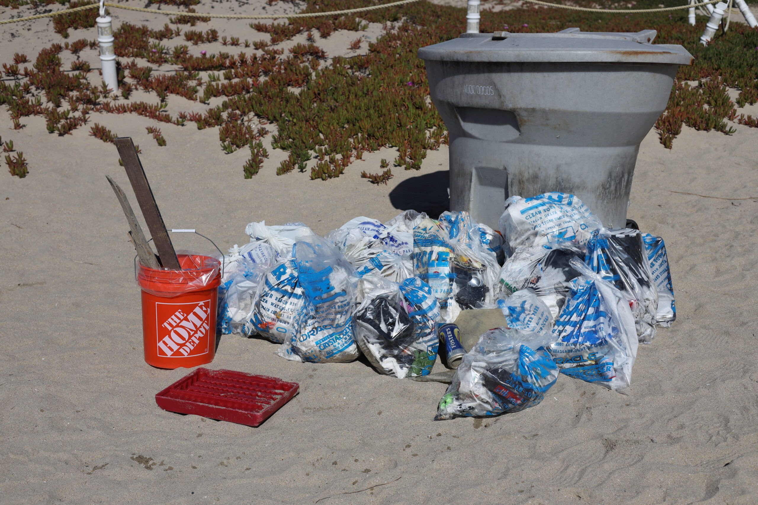 A trash can with bags of trash below it sitting on the sand.