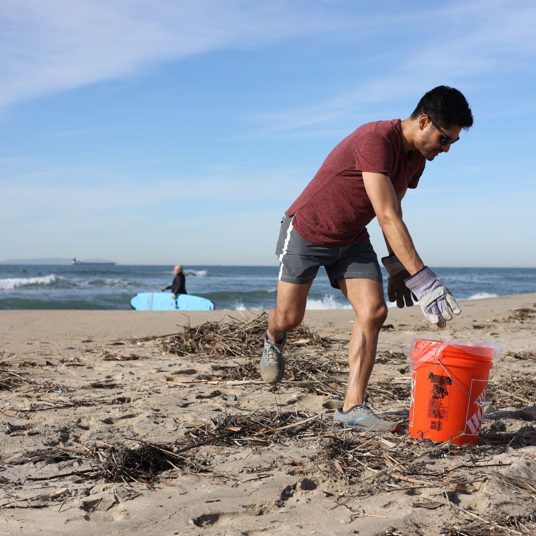 A man bends over to put trash in an orange bucket on the beach with blue hazy skies above.